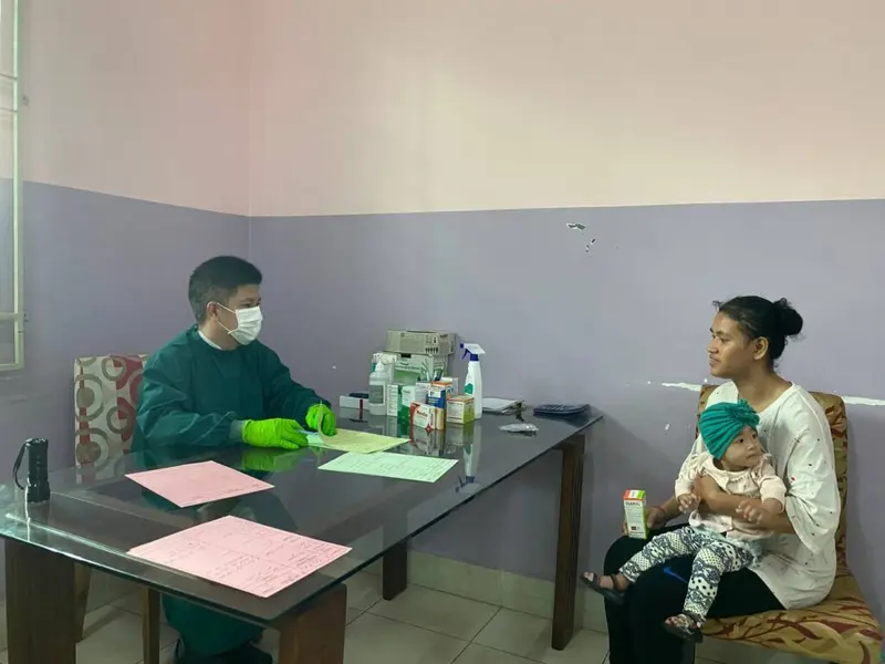 A doctor in green scrubs talks with a carer holding a small child in a clinic room.