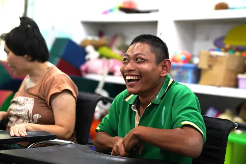 A young man in a green shirt laughs at a table in a bright classroom.