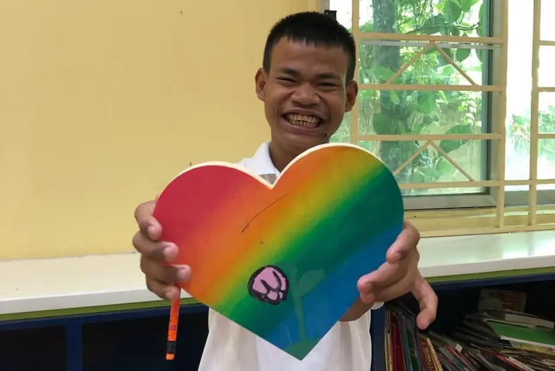A boy beams as he holds up a wooden heart painted in rainbow colors.