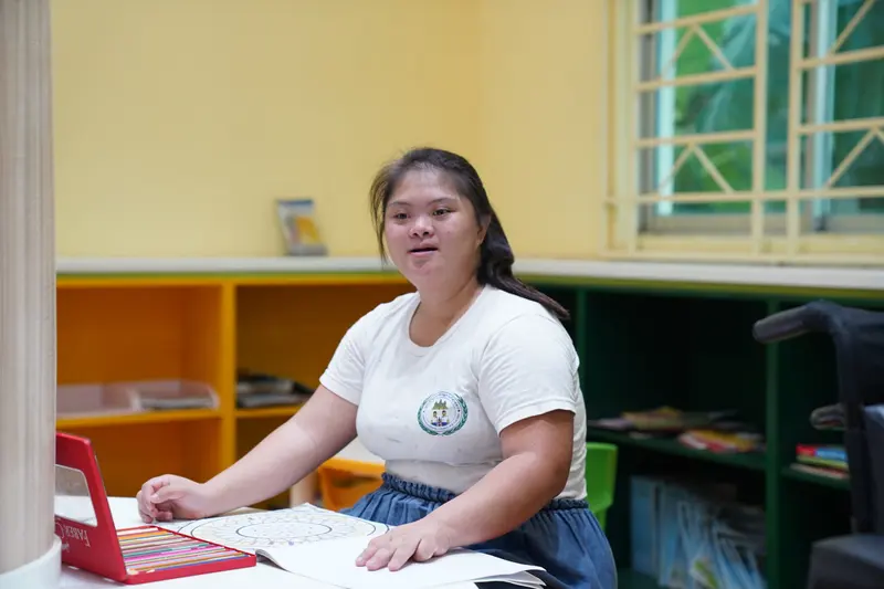 A young woman works at a desk in a sunny yellow classroom.